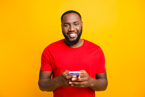 Photo of simple good cute handsome black man holding his phone with both, hands smiling at camera while isolated with yellow background