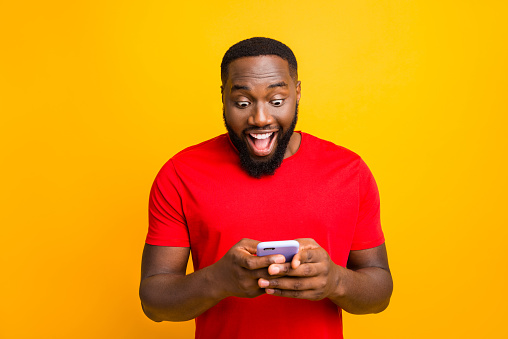 Photo of impressed black man watching something delightful on his phone wearing red t-shirt while isolated with yellow background