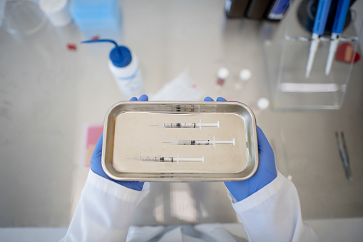 Three syringes are pictured on a sterile metal tray, which is held in the hands of someone wearing a lab coat and gloves. In the background are lab and research materials.