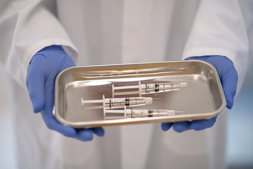 Three syringes are lined up on a metal tray, held in the hands of a professional wearing a lab coat and blue protective gloves.