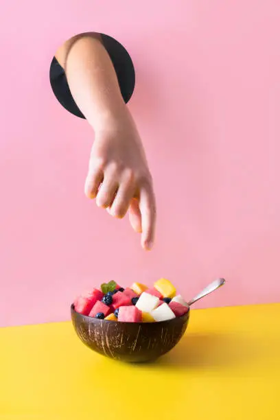 Photo of Childs hand through hole in pink paper reaches for fruit salad of watermelon in coconut bowl on yellow background. Food concept.