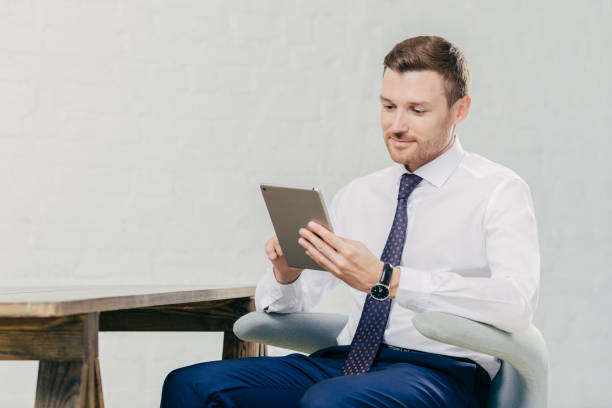 istock Prosperous handsome young businessman watches webinar online on modern tablet computer, dressed in formal clothes, sits at chair indoor. Young attractive male financier reads news on web page online 1169787170