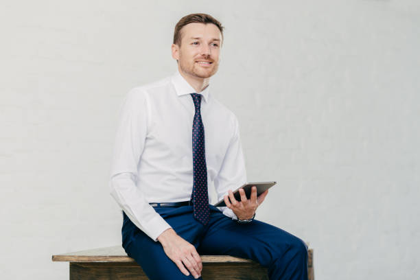 istock Thoughtful attractive businessman thinks about new creative idea, uses modern touch pad, dressed elegantly, sits on wooden table alone, poses against white wall. Prosperous male financier indoor 1169787200