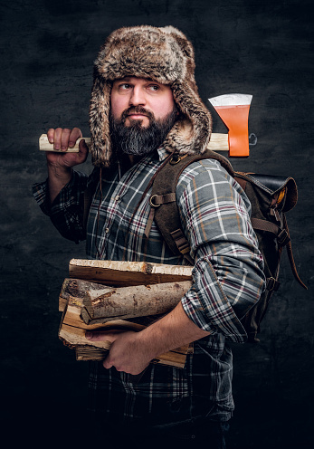 Retrato de un leñador barbudo con una mochila vestida con una camisa a cuadros y sombrero de trampero sosteniendo leña y hacha. photo