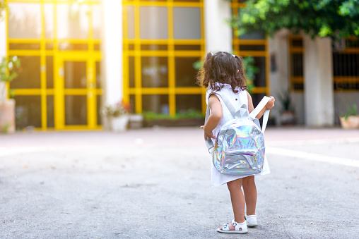 Back to school. Cute child girl with holographic backpack running and going to school with fun.Back, rear view of a toddler kid walking towards the preschool with the door in the background.