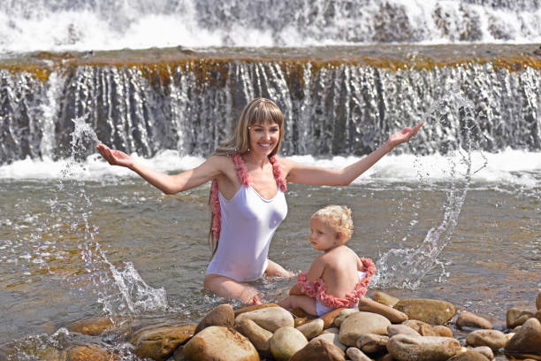 Mother with baby near waterfall. Young beautiful mother with her little daughter have fun near the waterfall. They are dressed in the same white swimwear.