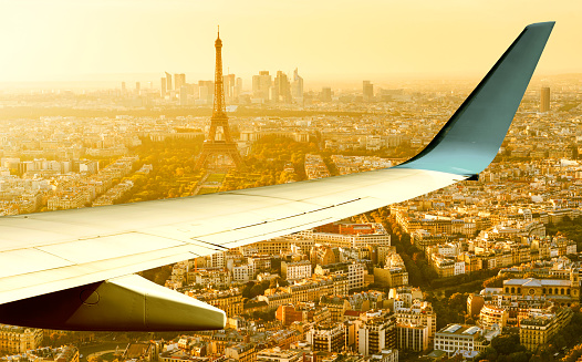 Plane flies above Paris in summer evening, France. Aerial panoramic view of Eiffel tower from airplane window. The plane's wing over streets of Paris at sunset. Flight and air travel to Paris.
