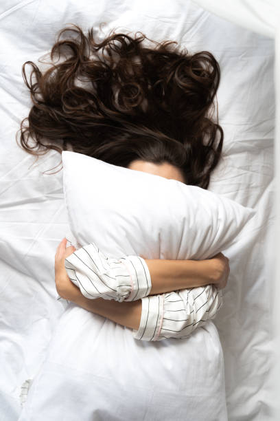 Young woman lying in her bed and hiding behind pillow