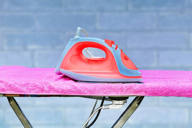 An ironing board with a power point is located in the living room. On the surface of the ironing board lies a clothes ironing iron connected to a household electrical outlet.
