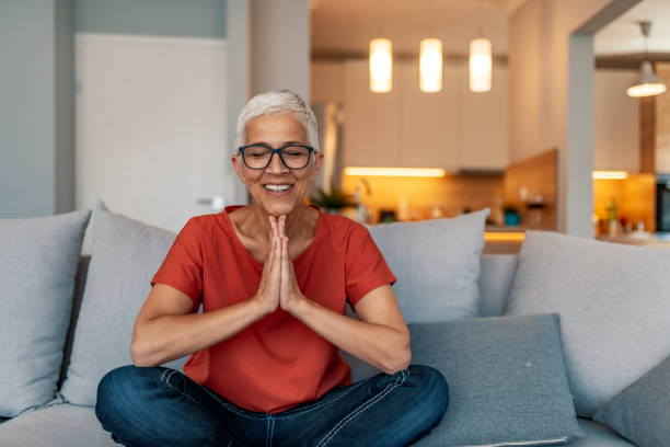 mature woman sitting on couch doing yoga exercises at home - om symbol fotos imagens e fotografias de stock