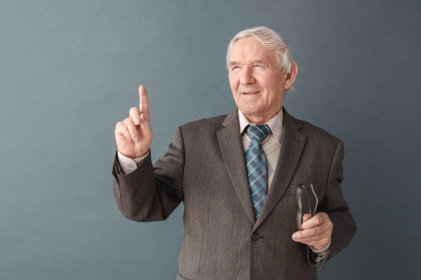 istock Senior man teacher studio standing isolated on gray holding glasses looking aside illuminated 1169834938