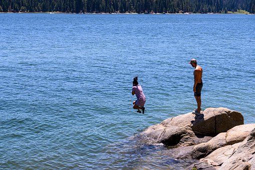 One young female Latino adult jumping off the rocky shore, of remote mountain lake, with male Latino adult waiting for his turn.

Taken at Shaver Lake, California, USA