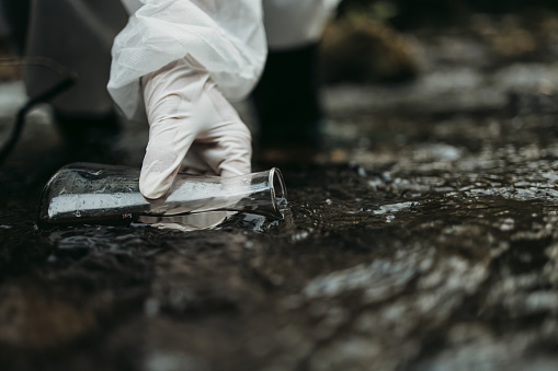 Close up shot of scientist biologist and researcher in protective suit taking water samples from polluted river.