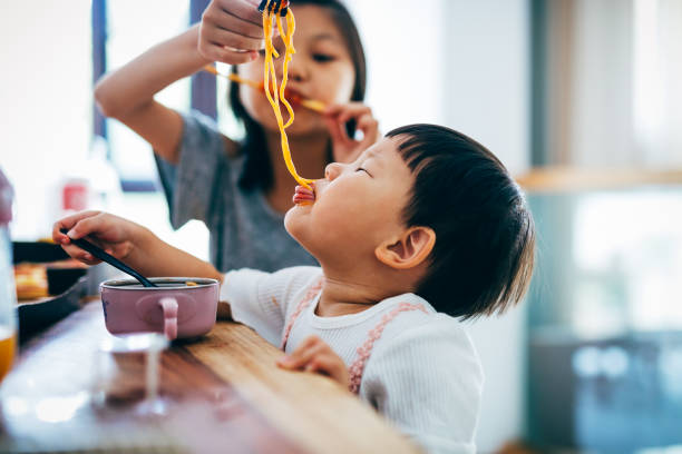 Little Asian girls and her sister eating noodles at home. Eating, Baby - Human Age, Messy, Toddler, Pasta, Asian and Indian Ethnicities, Chinese Ethnicity, Girls, Shanghai kids eating noodles stock pictures, royalty-free photos & images