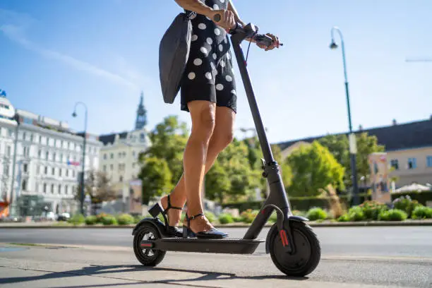 legs of 45 years old business woman in summer dress with bag on electric scooter on cycle path through historical city center in vienna on sunny day, waist down, background blurred