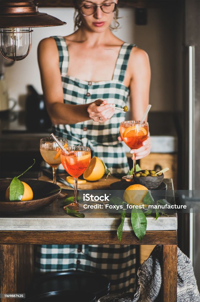 Young woman holding Spritz cocktail and olive Young woman in checkered dress holding Spritz aperitif drink with orange in glass and olive in hands, kitchen at background. Summer refreshing drink concept Cocktail Stock Photo