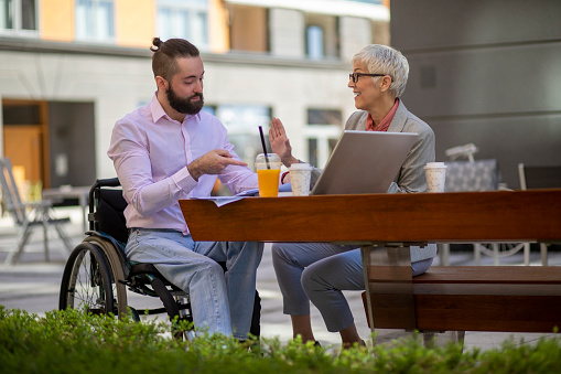Young man in the wheelchair with the laptop having a business meeting with a mature gray haired woman in a public park.  The woman is sitting on a bench.