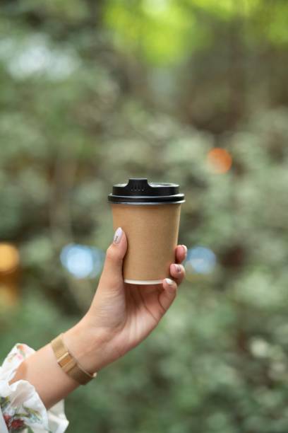 istock Young woman hand holding brown paper coffee cup with blurred tree background 1169941143