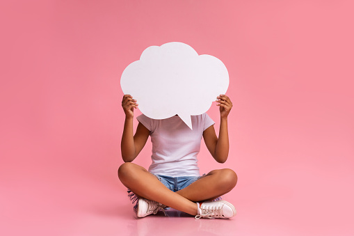 Empty speech bubble at black teenager hands. Girl sitting with crossed legs at pink studio background