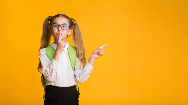 istock Elementary Student Putting Finger To Lips Pointing Aside, Yellow Background 1169957885