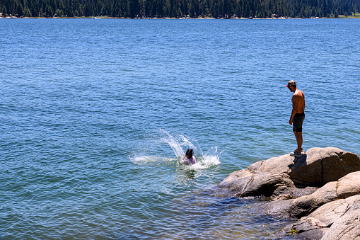 One young female Latino adult jumping off the rocky shore, of remote mountain lake, with male Latino adult waiting for his turn.

Taken at Shaver Lake, California, USA