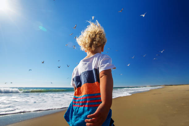 A teenage boy walks alone on the beach during sunset. The boy's expression is contemplative, reflecting the peaceful and introspective atmosphere of the moment. The serene setting, with the calming sounds of the ocean and the warm colors of the sunset, captures a quiet and reflective slice of time.
