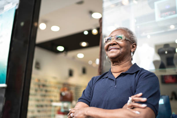 senior business owner standing in front of her store looking away with arms crossed - owner boutique store retail imagens e fotografias de stock