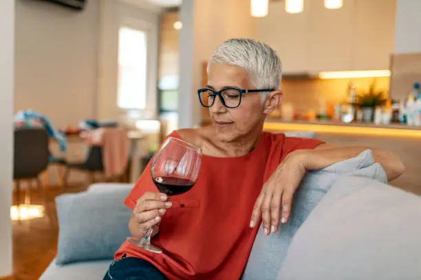Photo of mature sad woman sitting on sofa with one glass of red wine waiting for somebody (man) - loneliness, quarrel, celebration Valentine Day alone concept. Lonely mature woman holding glass of alcoholic drink while sitting on sofa at home during the day.