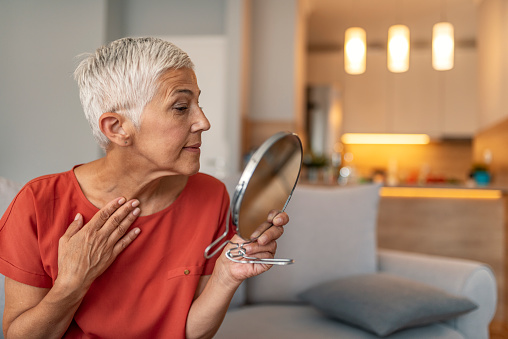 Beautiful short hair elderly woman holding mirror and applying face cream at home. Attractive gray hair senior woman looking at herself in mirror while sitting on sofa during the day.