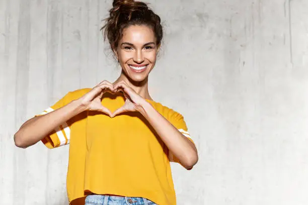 Portrait of beautiful smiling brunette woman wearing mock-up yellow shirt and showing heart with hands. Copy space in right side.