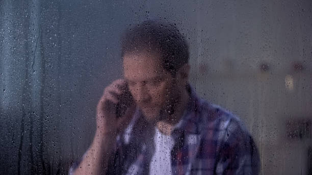 istock Middle-aged male talking phone with family behind rainy window missing relatives 1169983354