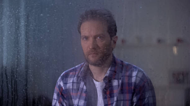 istock Lonely man sitting behind rainy window and looking at camera, depression concept 1169983365