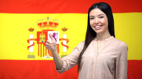 Smiling Asian woman showing thumbs-up gesture against Spanish flag background