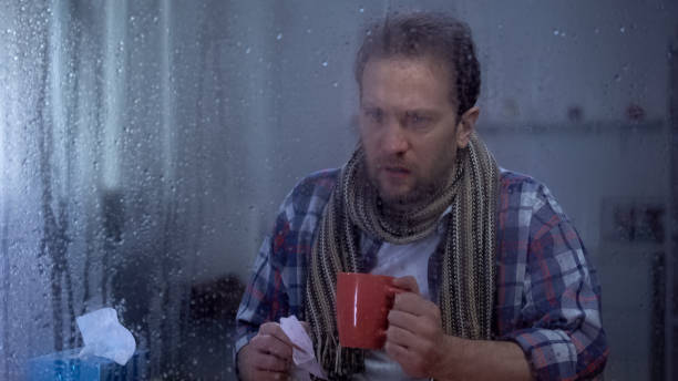 istock Sick man with handkerchief drinking hot tea at home behind rainy window, flu 1169984730