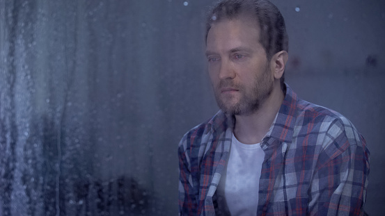 Sad man looking through window during rainy weather, thinking about life problem