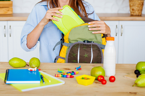 Healthy food concept. Closeup of female hands packing lunch box into a kid's rucksack in the kitchen. Selective focus.