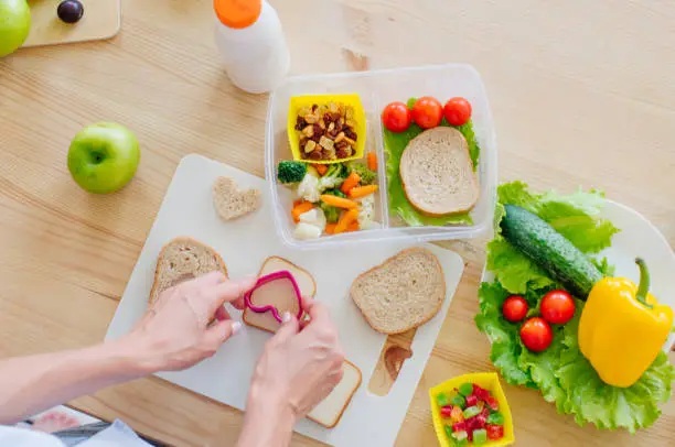 Healthy food concept. Closeup of female hands preparing sandwich for lunch box filled with bread, fresh vegetables, fruits and snack on the wooden table. Top view, selective focus.