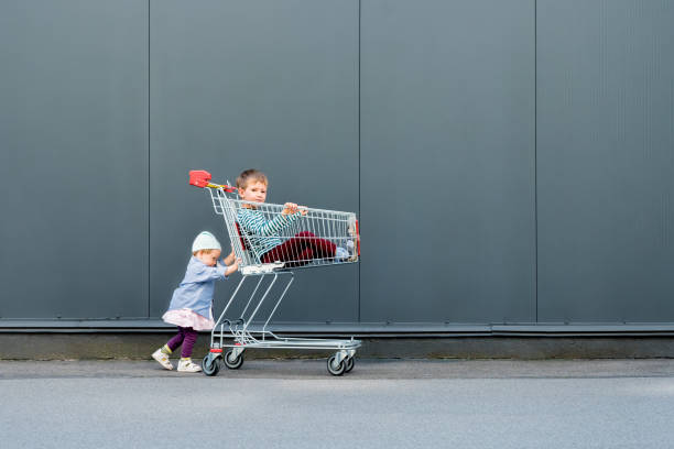 dois hipsters na moda com o carro de compra perto do supermercado, alameda de compra. duas crianças bonitos que esperam pais ao ar livre. fundo feliz do conceito da infância. menina que joga com seu irmão - carrinho de criança - fotografias e filmes do acervo