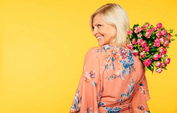 Photo of Holiday event. Mother's day. St. Valentines day. Birthday. Portrait of happy cute lovely senior woman in party colorful clothes with large bouquet of flowers on yellow background