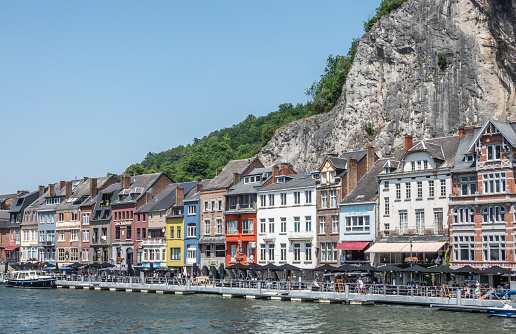 Dinant, Belgium - June 26, 2019: Row of colorful business facades just past Charles de Gaulle bridge on norht right bank of Meuse River under blue sky, Gray Citadell cliff in back.