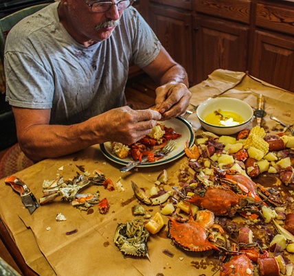 Older Caucasian man with gray moustache and unusually large hands picking seafood with a utility knife at a Cajun seafood boil including crabs, crawfish shrimp potatoes and corn served traditionally on brown paper