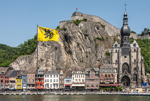 Dinant, Belgium - June 26, 2019: Notre Dame church and Citadelle from other river Meuse bank with businesses on right north bank, under blue sky. Green foliage. Flag of Flanders.