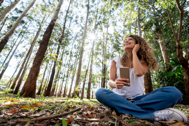 happy woman listening to music outdoors on her cell phone - tree stream forest woods imagens e fotografias de stock