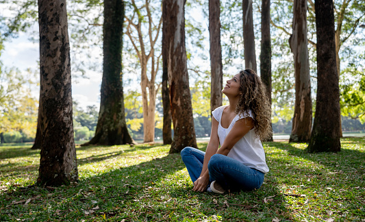 Happy young woman relaxing at the park sitting on the floor and looking at the trees while smiling
