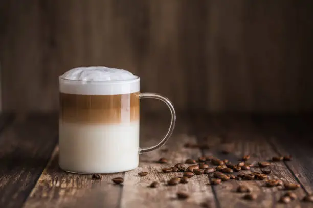 Cafe latte macchiato layered coffee in a see through glass coffee cup. The cup is on a wooden background with coffee beans on the table next to the cup.