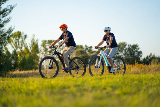 active senior couple cycling on mountain bikes through rural landscape happy senior couple cycling through nature reserve on their modern electric bicyles mountain bikes on sunny summer day electric bicycle photos stock pictures, royalty-free photos & images