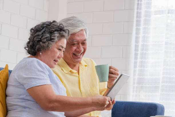 asian senior couple using table compute and drink coffee sitting at sofa in living room at home.senior with technology lfiestyle.aging at home concept. - ipad senior adult facebook sofa imagens e fotografias de stock