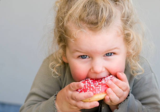 Little girl eating jelly-glazed donut with sprinkles Young girl aged 2 to 3 years eating donut with pink icing overweight child stock pictures, royalty-free photos & images