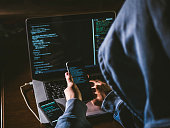 anonymous person in the hood sitting in front of computer working with laptop and mobile phone