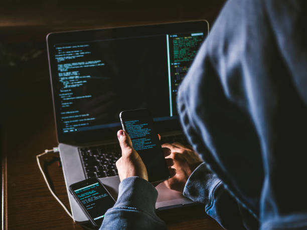 анонимный человек в капюшоне, сидящий перед компьютером, работающим с ноутбуком и мобильным телефоном - хакер стоковые фото и изображения